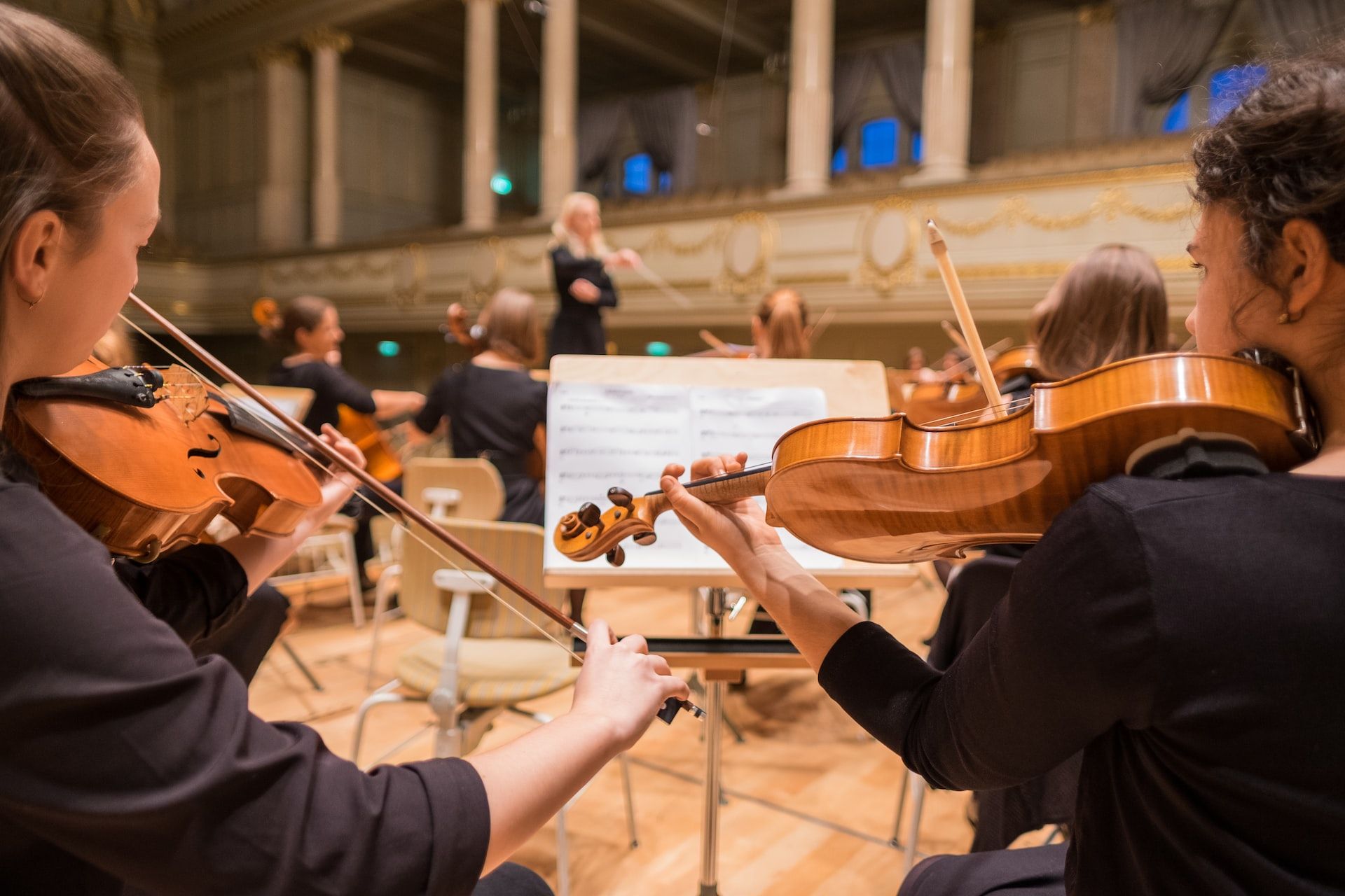 Two violinists in an orchestra playing from one music stand, conductor in the background